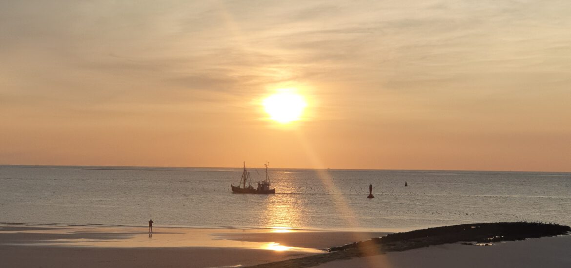 Mein Happy Place auf dem Bild sieht man einen Sonnenuntergang am Strand. Vereinzelte Schleierwolken befinden sich am Himmel. Ein kleines Fischerboot dümpelt auf dem ruhigen Meer vor sich dahin. Es herrscht eine friedliche Stimmung.