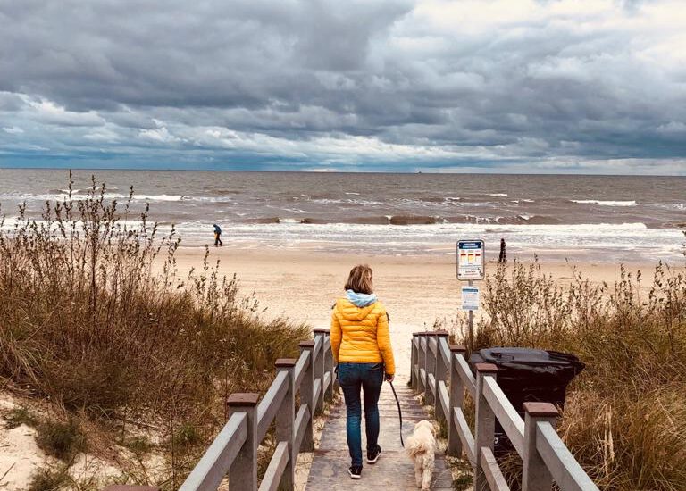 Wann öffnen die Campingplätze? Das Bild zeigt Andrea und ihren Hund Polly von hinten, wie sie über einen Steg zum Strand von Usedom gehen. Im Hintergrund sieht man tiefe Wolken und eine starke Brandung.